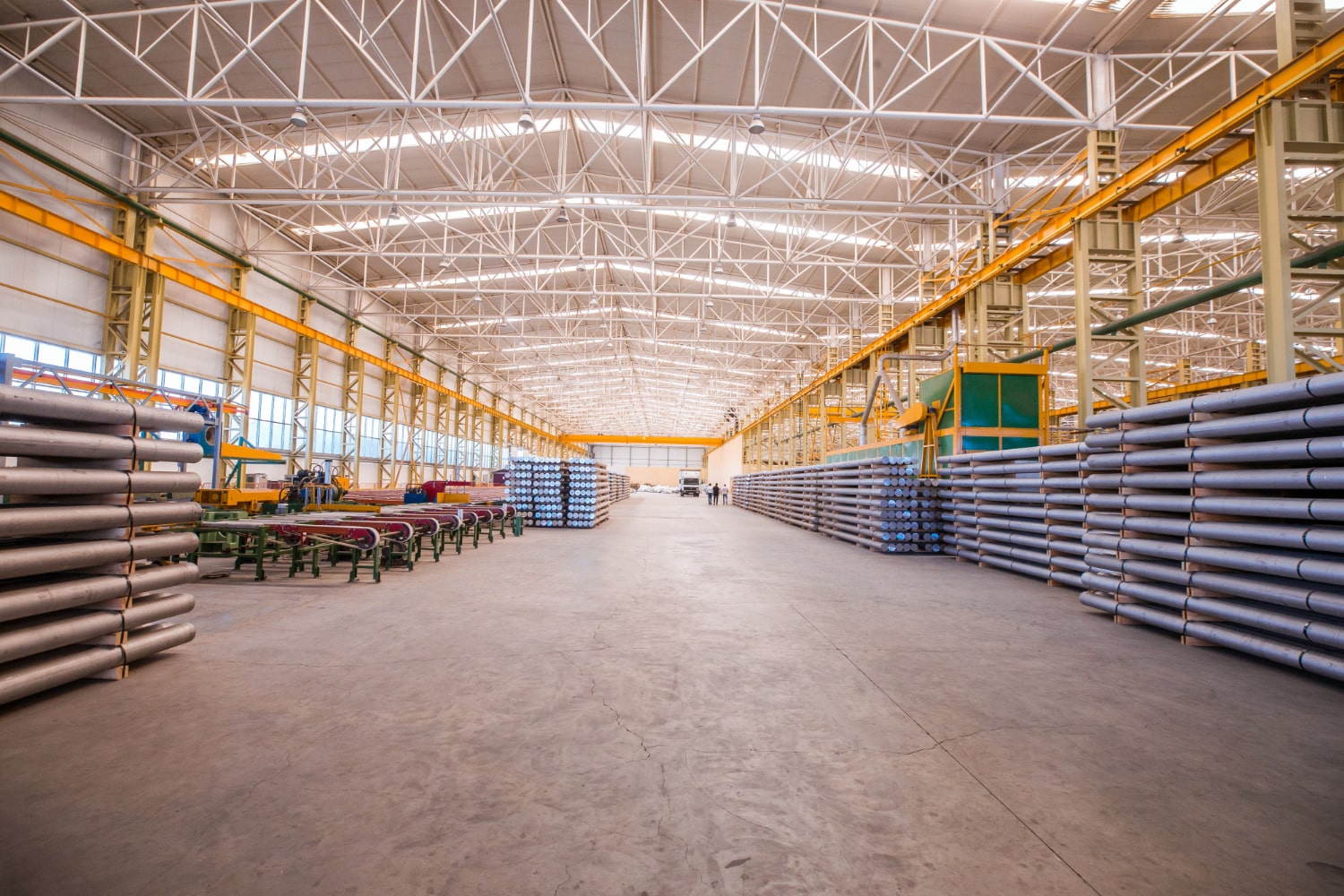 A spacious industrial warehouse with stacks of metal pipes, a high white truss ceiling, and bright warehouse lighting illuminating the space.
