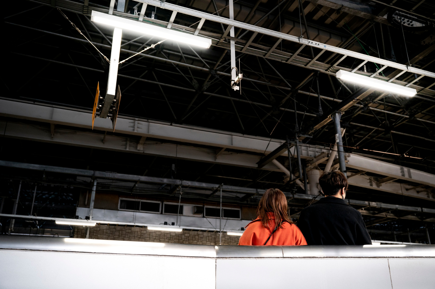 Two people, seen from behind, stand under bright lights in an industrial-looking indoor space.