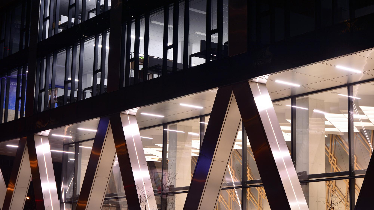 Modern office building at night with large glass windows and illuminated diagonal steel beams, showcasing sophisticated commercial lighting that enhances its striking architectural features.