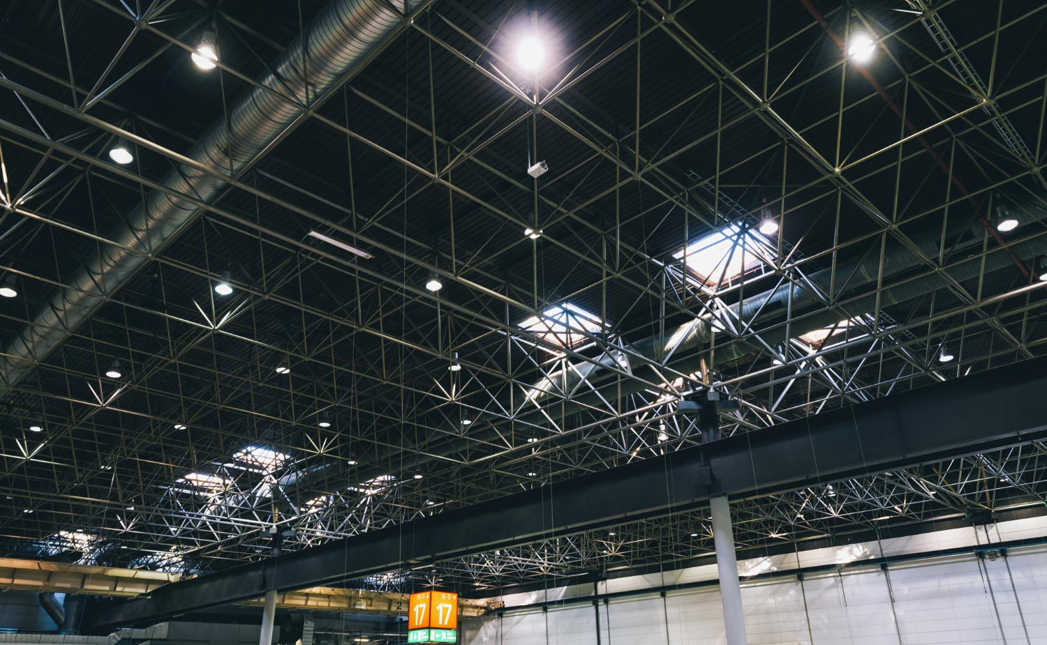 Industrial ceiling with metal beams, lights, and ducts in a large indoor space—possibly an airport or warehouse—showcasing a comprehensive lighting inventory integrated throughout the structure.